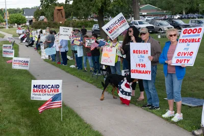 Community members rally along a sidewalk with signs reading "Medicaid Saves Lives" and "Good Trouble" at the Good Trouble rally in Brookings, South Dakota.